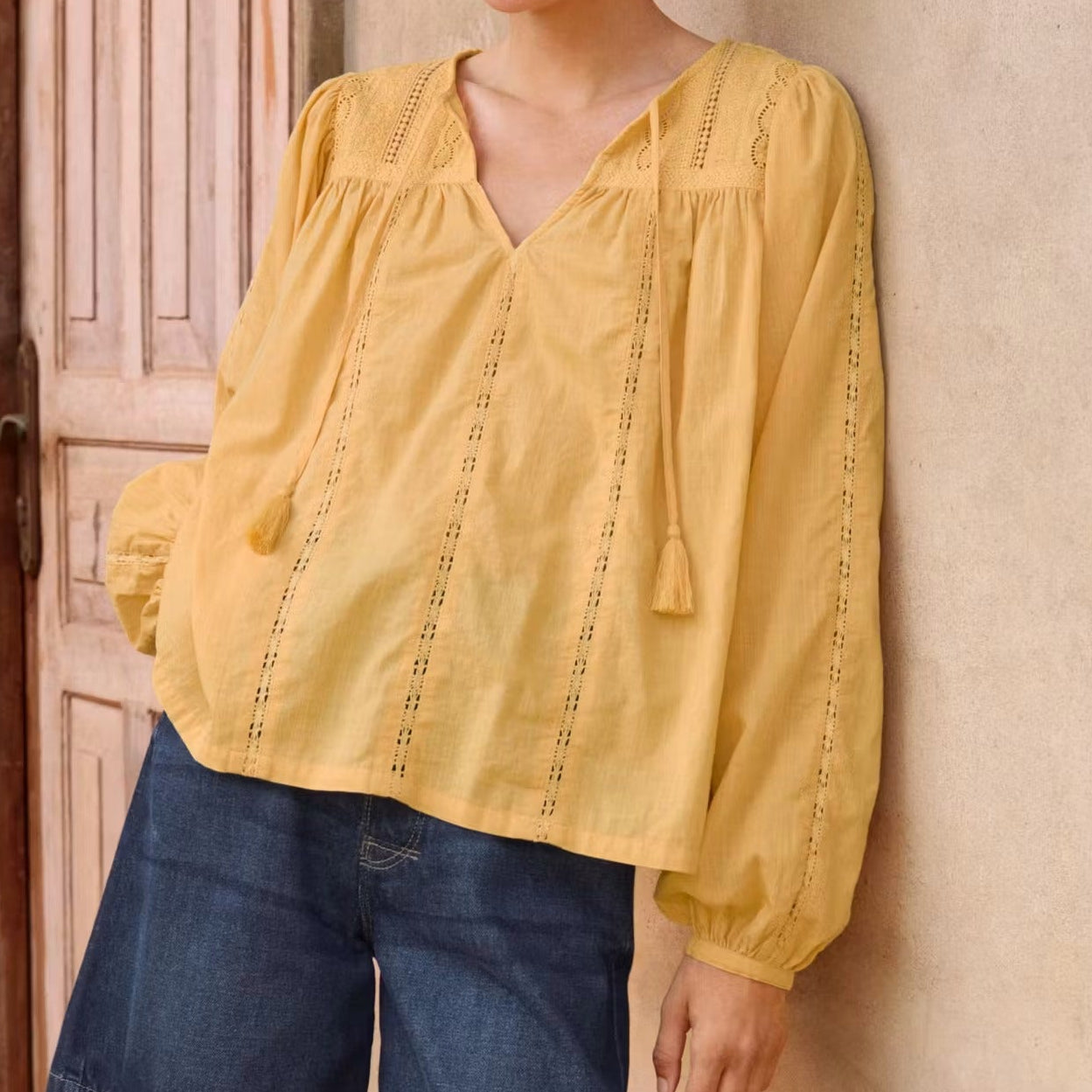 Woman wearing a yellow blouse and blue shorts standing against a wall.