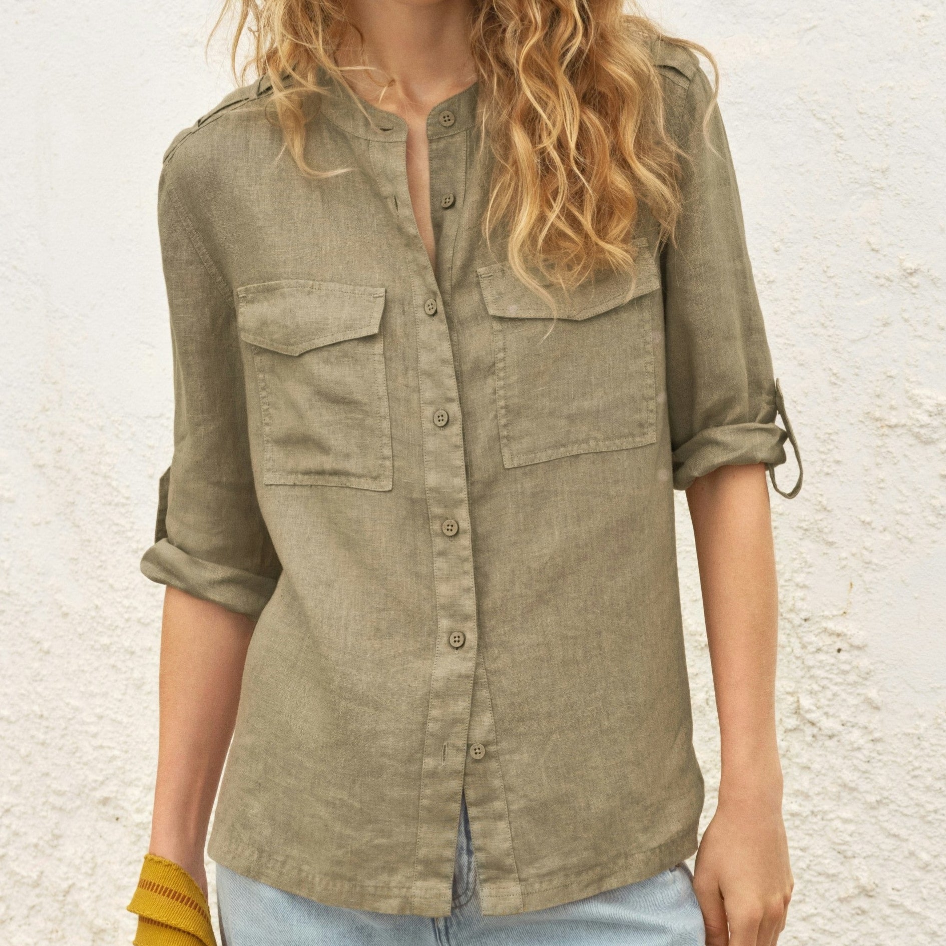 Woman wearing an olive green shirt with rolled-up sleeves against a textured wall.