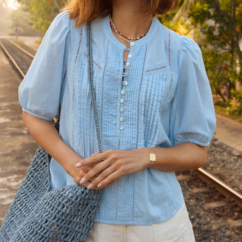 Woman in a light blue shirt and beige shorts holding a blue bag on a path with greenery.